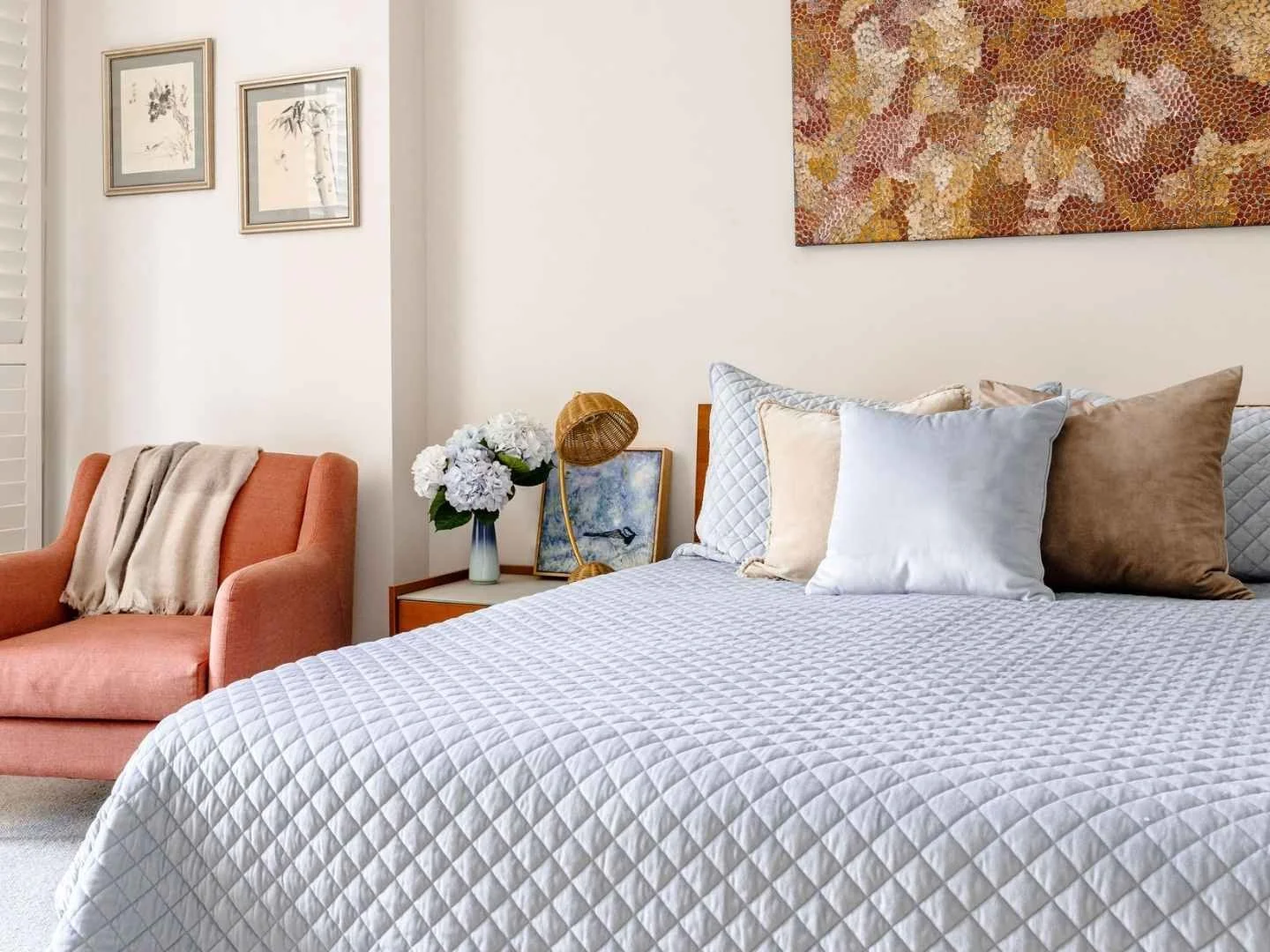 Bedroom interior with a light blue quilted bed, layered neutral cushions, coral armchair, framed artwork and bedside styling.