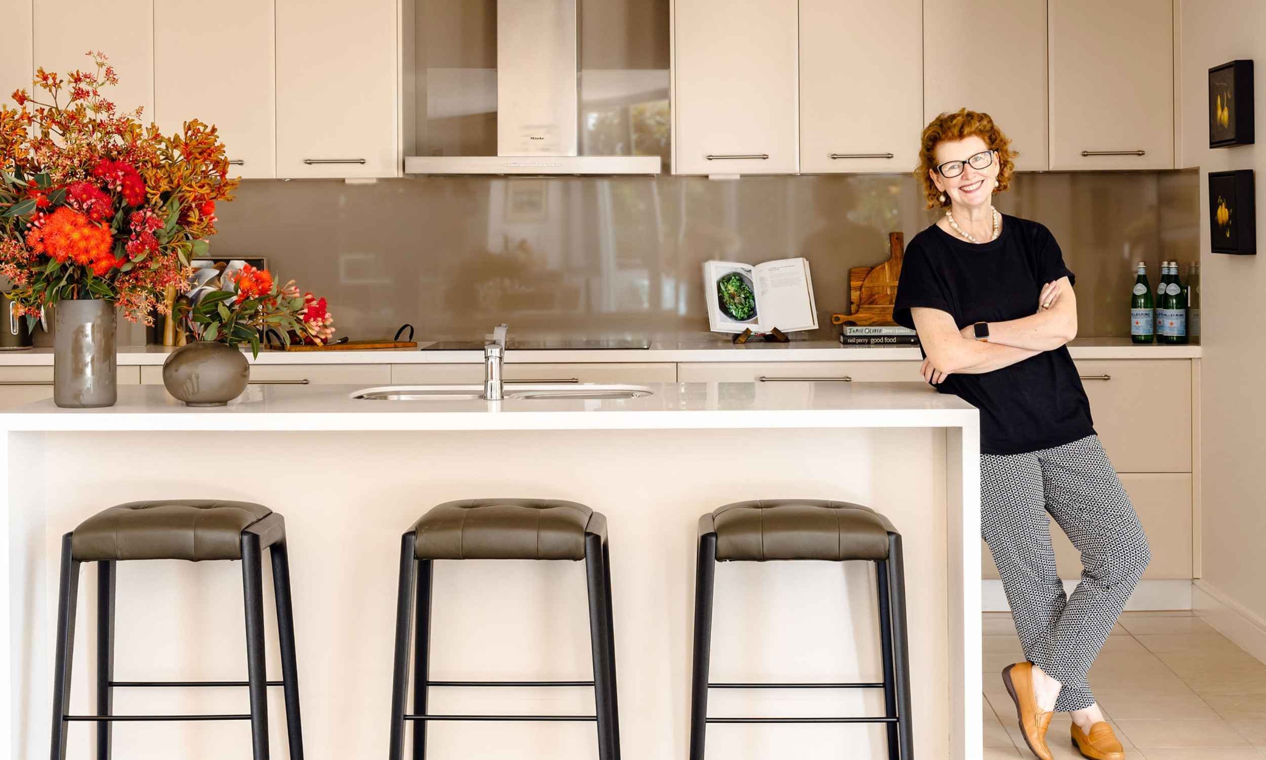 Kate Kerin standing in a contemporary Greenwich kitchen with a white island bench, neutral cabinetry and large floral arrangements.
