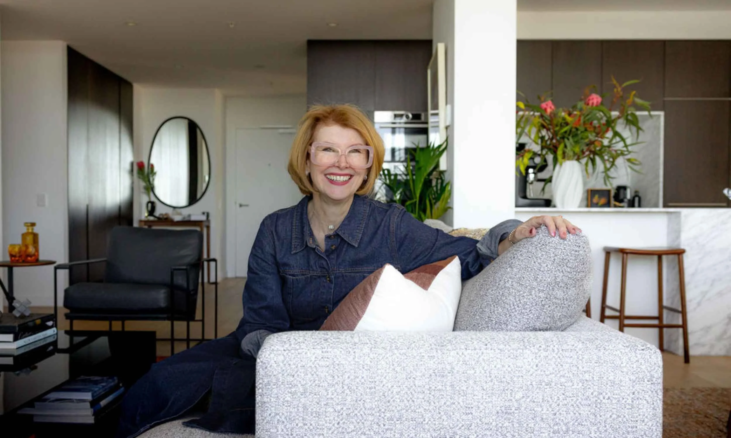 Kate Kerin smiling whilst sitting on a textured sofa in a contemporary living room, with a marble kitchen and floral arrangement in the background.