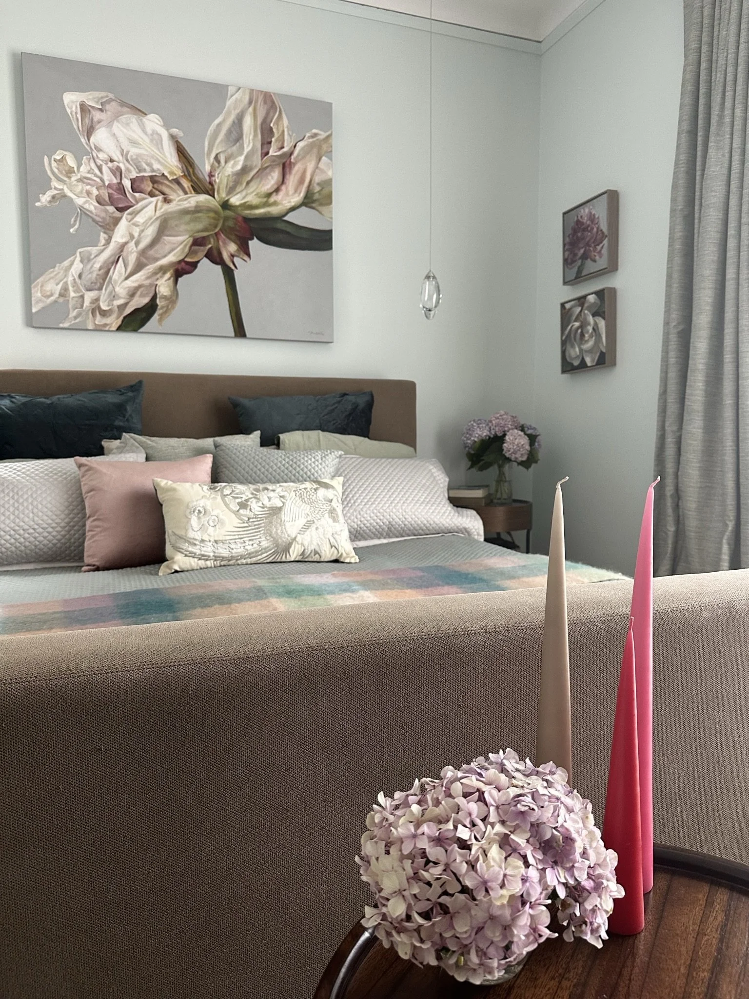 This image is a close up of a bedroom, with a painting above the bed of an overblown peony. In the foreground are pink candles and a small vase of pink hydrangeas.