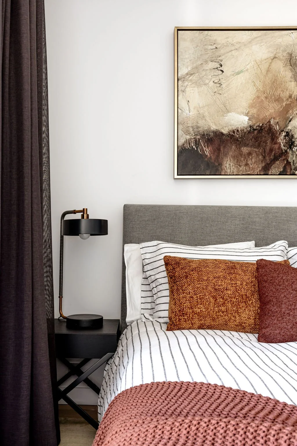 A masculine bedroom with rust and burgundy cushions on the bed. A pretty artwork above the bed and black curtains.