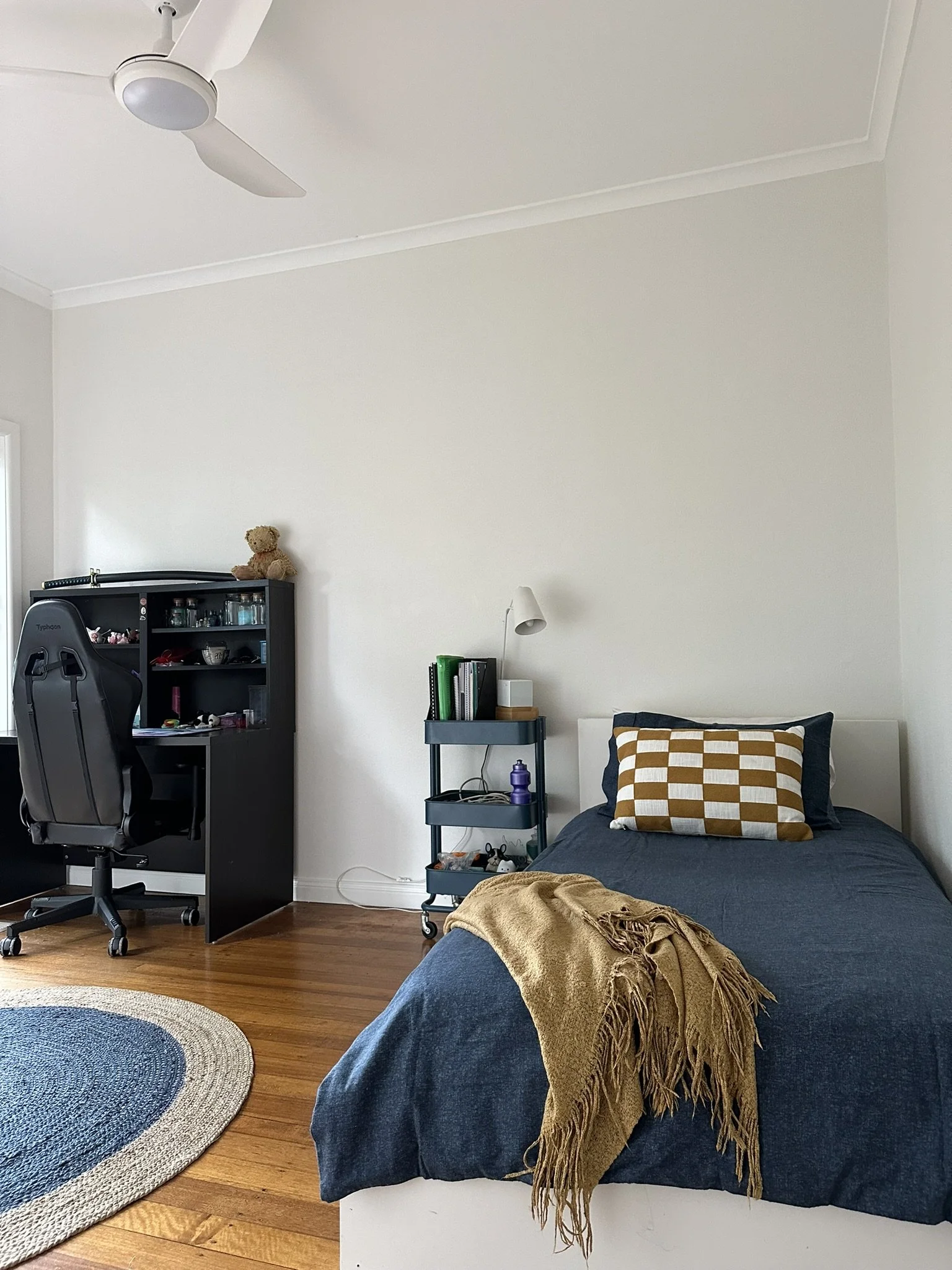 A stylish boy's bedroom with desk in the background and a single bed. The bed has a navy blue quilt and mustard coloured throw across the bottom, with a mustard-and-white checked cushion near the pillow. A ceiling fan is in the centre of the ceiling.