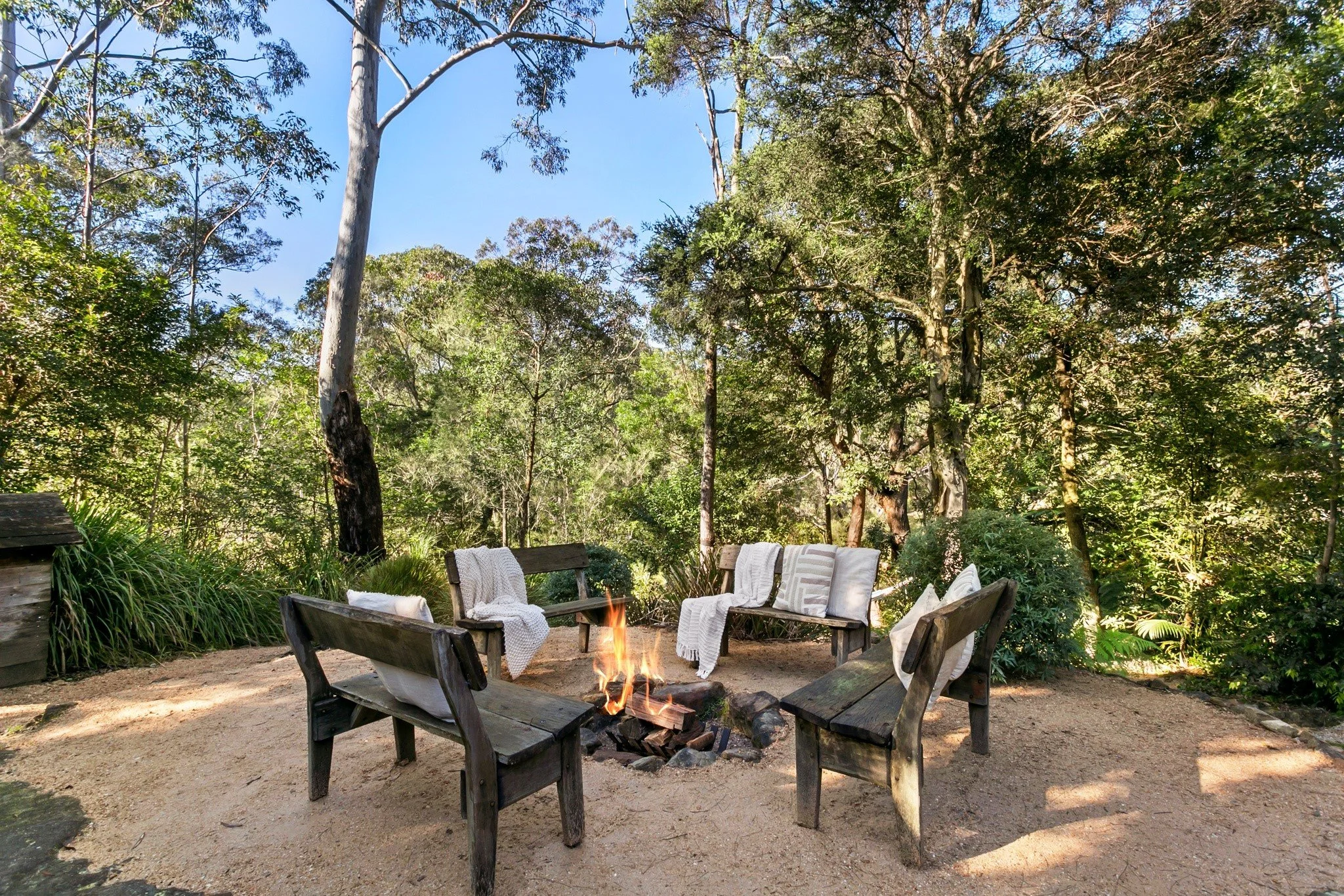 An outdoor fire pit with bushland all around it, set with wooden chairs and benches.