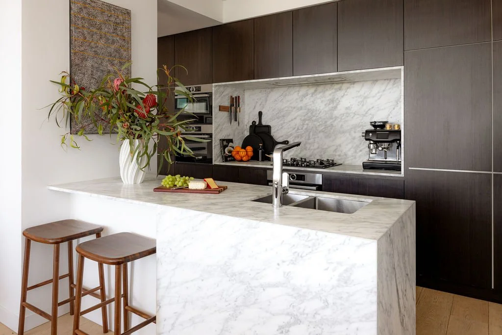 Galley kitchen with vase of flowers, artwork above island and cabinetry in the background. Wooden stools under the bench.