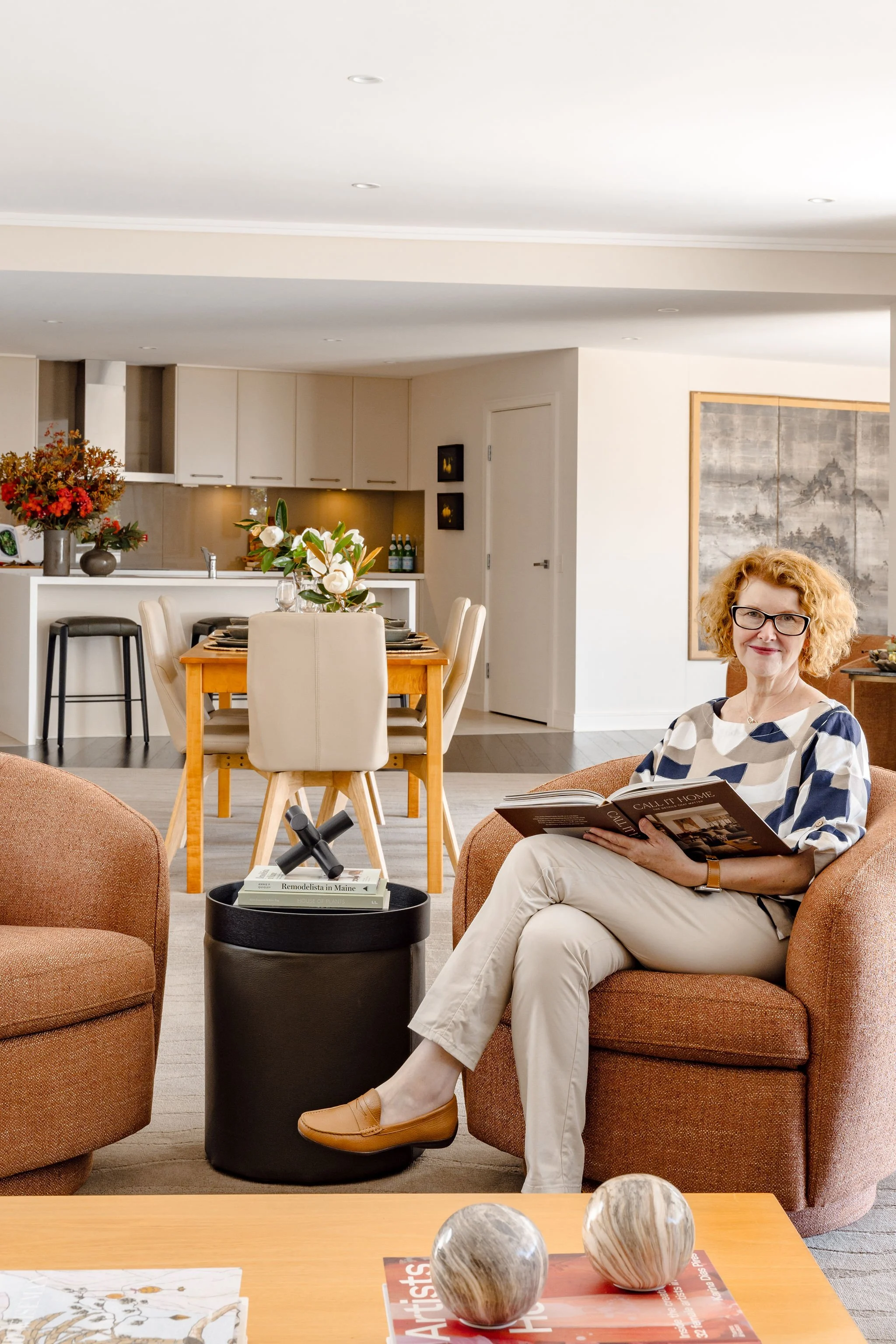 Kate sits with her legs crossed, in front of a dining area and kitchen in the background. She is waring glasses and a neutral, geometric top and is reading a book.