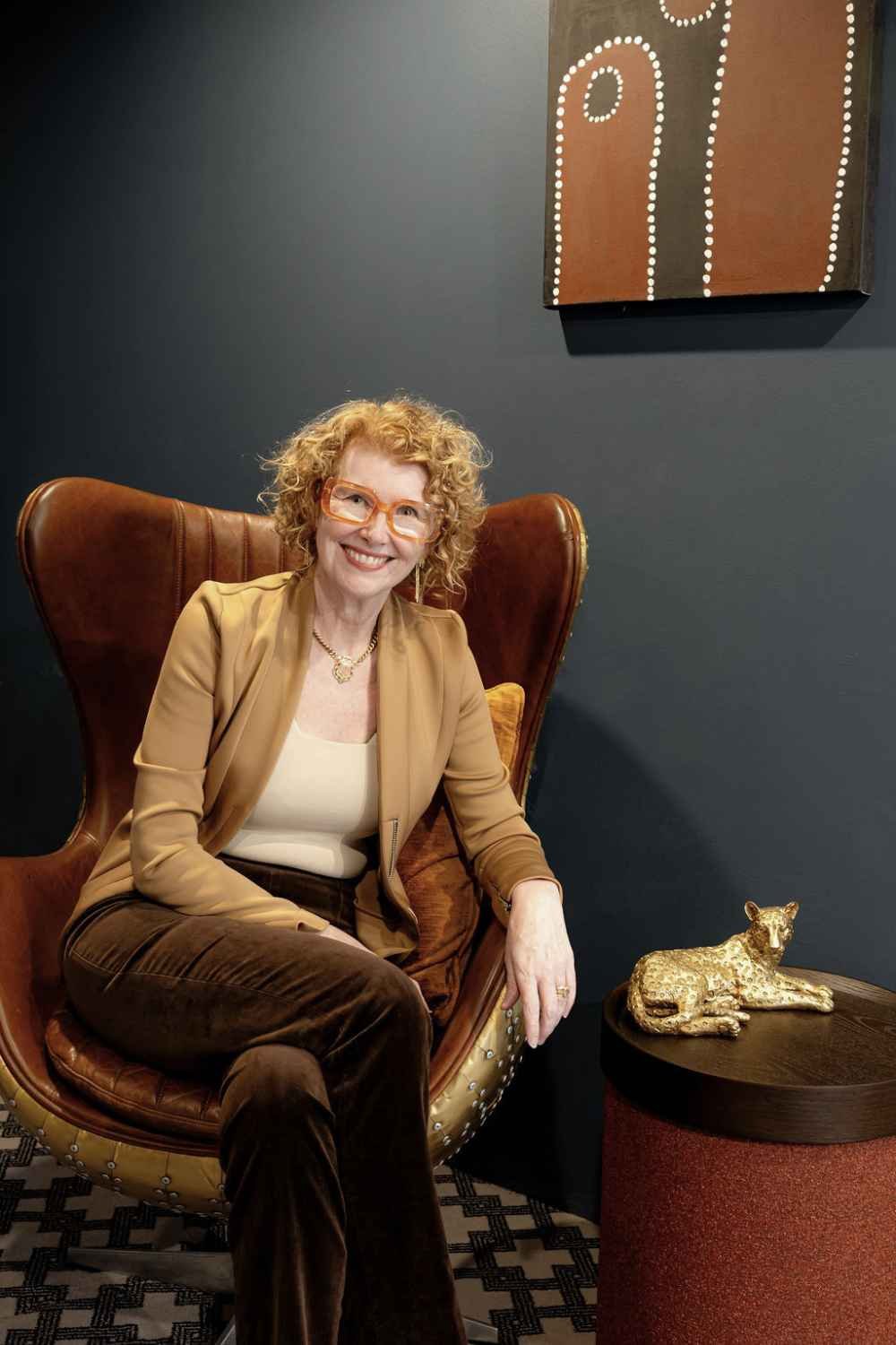 Kate Kerin seated in a brown leather armchair beside a small table with a gold cat sculpture and artwork on a dark wall.