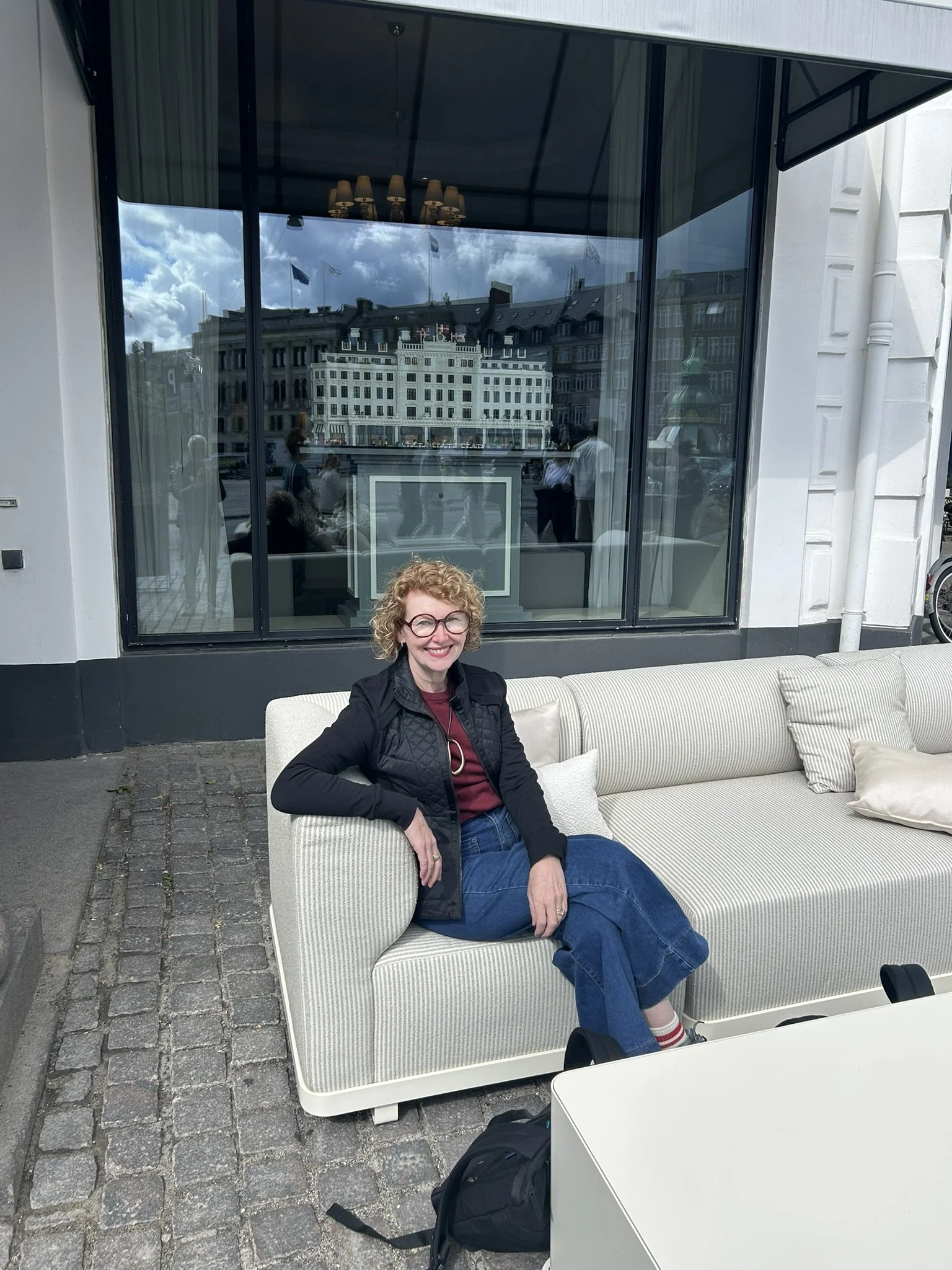Kate sits on an outdoor sofa, in Copenhagen. She's wearing jeans and a black jacket, plus round glasses. Behind her is a model of a beautiful building.