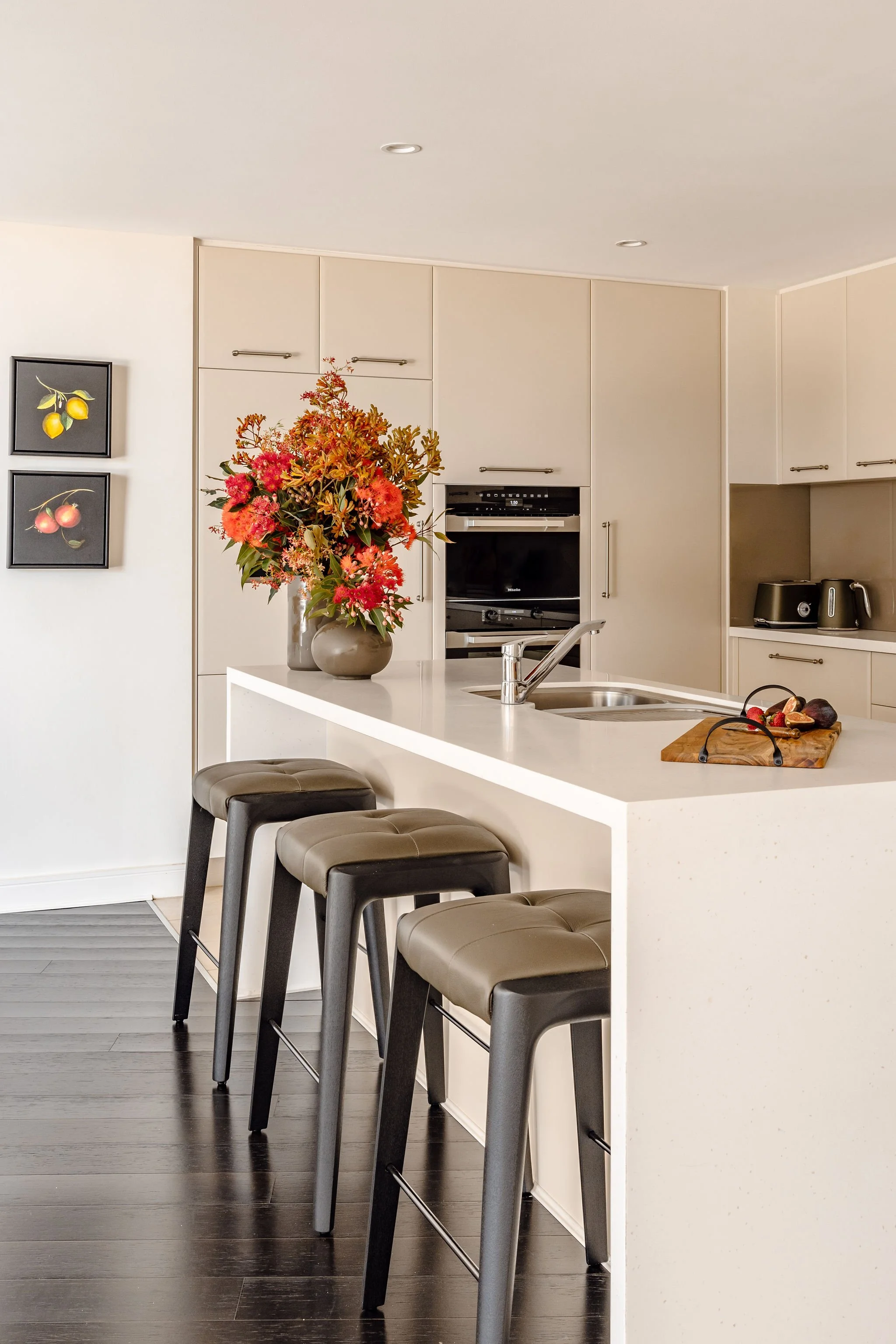 A modern kitchen with a large bunch of native flowers on the island. In the background are two paintings of citrus fruit, and under the island are three olive coloured leather stools.