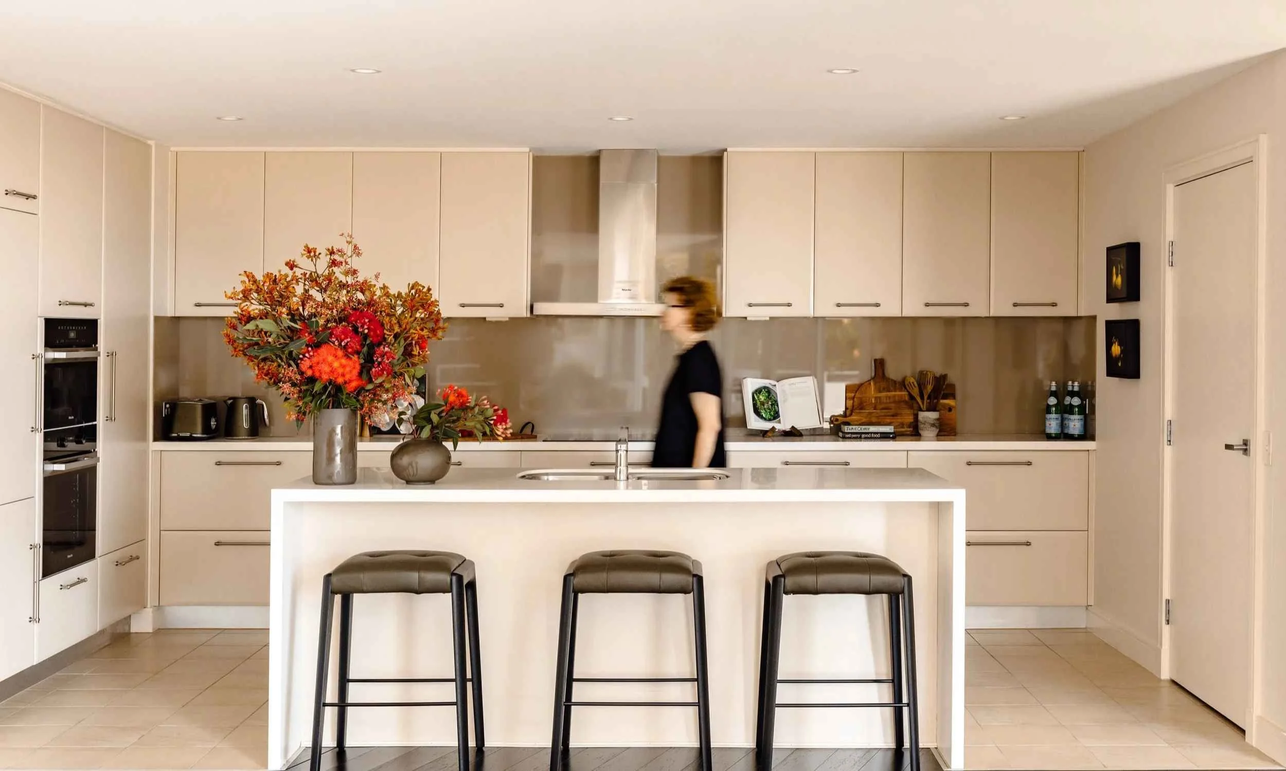 Kate Kerin walking through a neutral Greenwich kitchen with white cabinetry, island bench and large floral arrangement.
