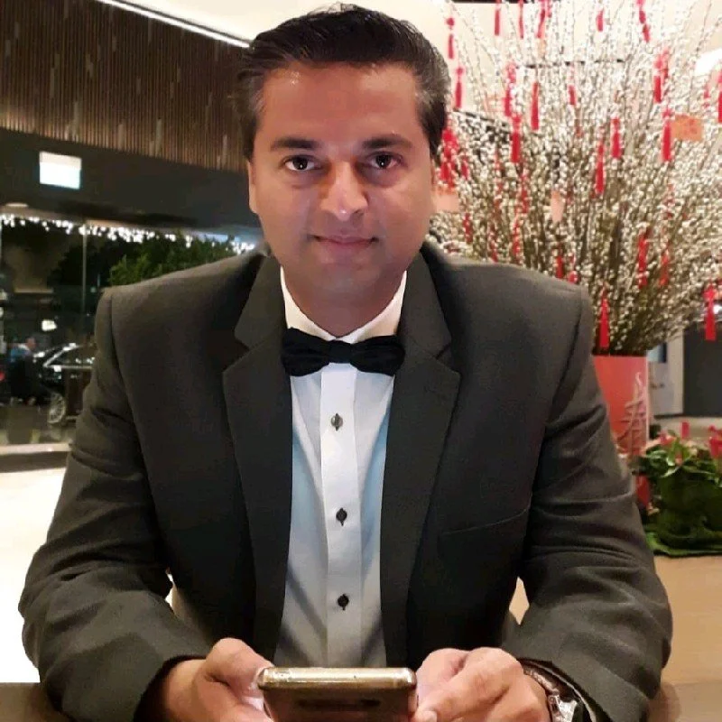A man in a black tuxedo and bow tie sitting at a table, holding a smartphone, with a decorative floral arrangement in the background.