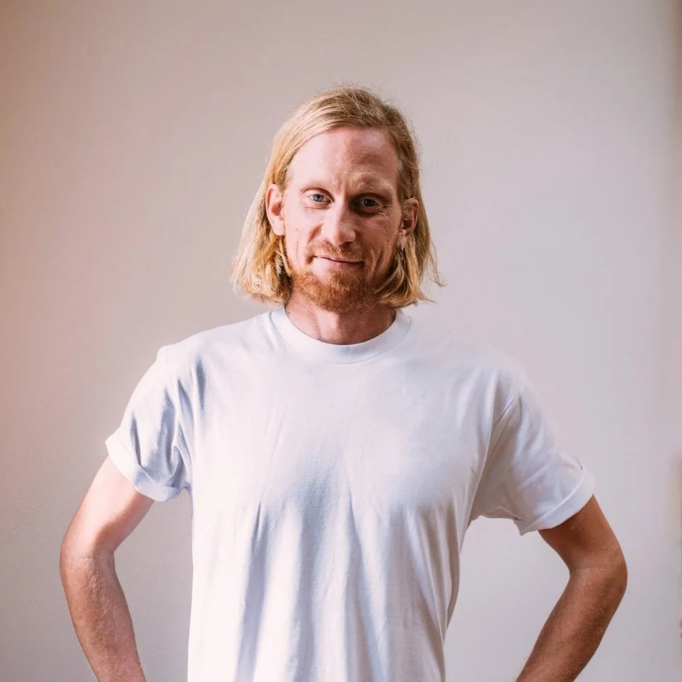 A man with long blonde hair, a beard, wearing a white T-shirt, standing with hands on hips against a neutral background.