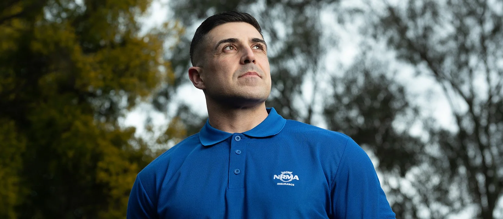 A man in a blue polo shirt with an 'NRMA Insurance' logo looking off into the distance outdoors, with blurred trees in the background.