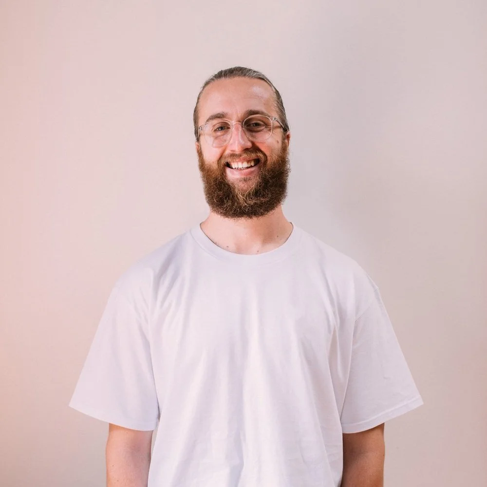 A man with a beard and glasses smiling at the camera, wearing a white t-shirt, against a plain background.