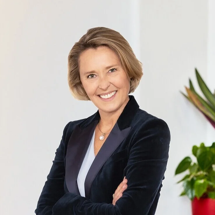 A woman with blonde hair smiling and standing with her arms crossed, wearing a dark blazer and white top, in front of a plain light-colored wall with a green plant in the background.