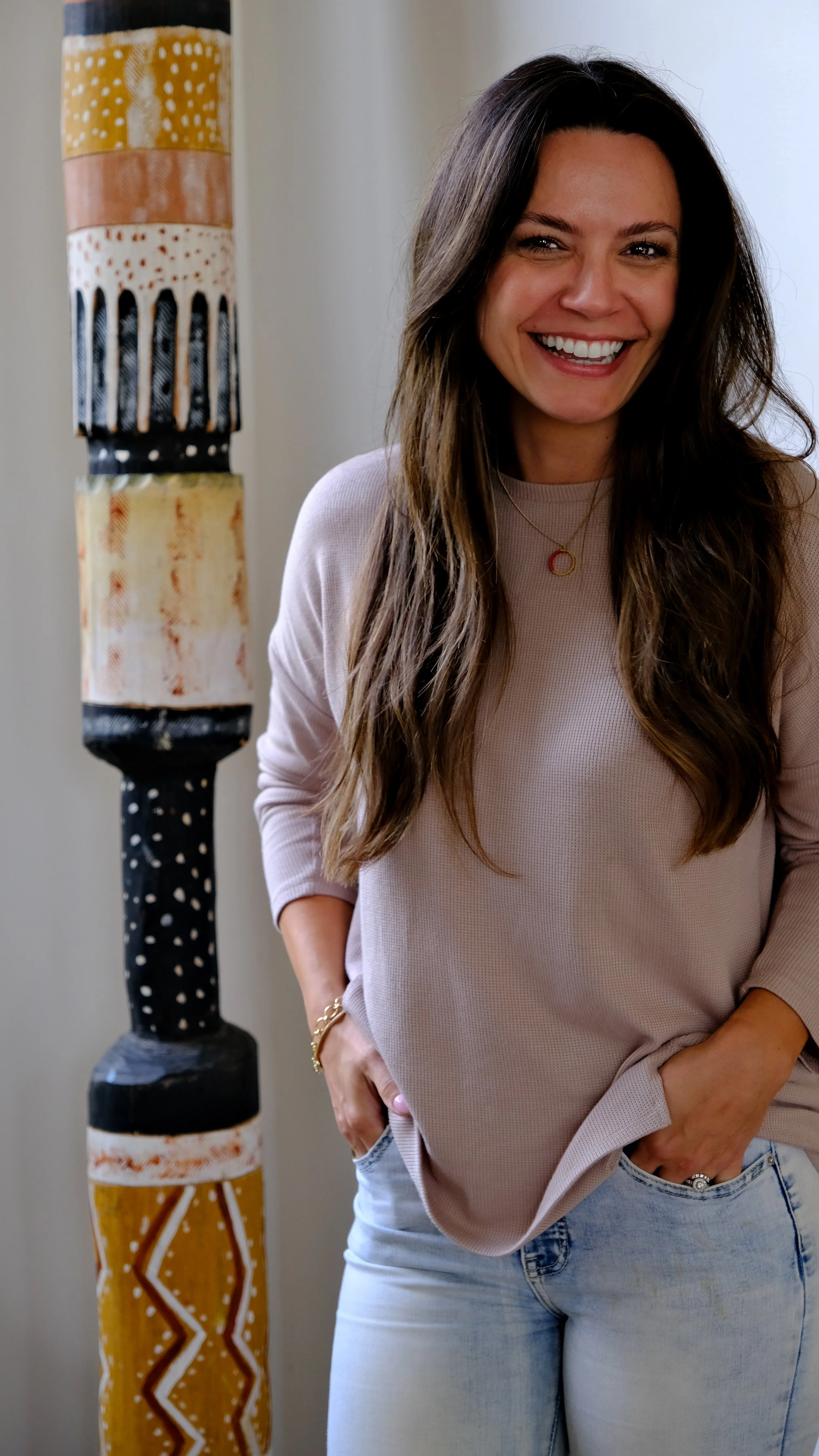 A woman with long wavy brown hair, smiling, wearing a light pink long-sleeve shirt and light-wash jeans, standing next to a tall, colorful, painted sculpture.