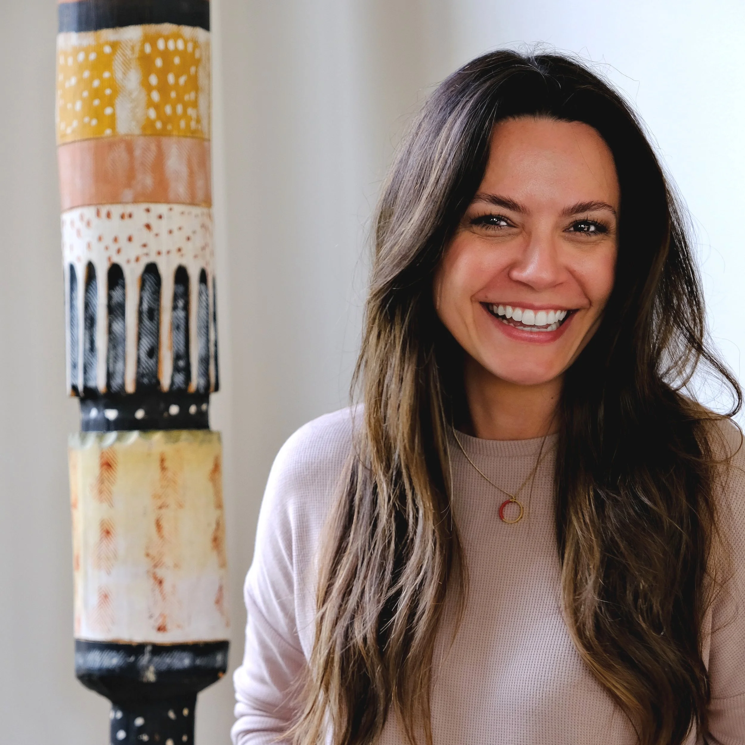 A woman with long wavy brown hair and a big white smile, wearing a light pink top and a necklace with a red circle, standing next to a colorful striped lamp.