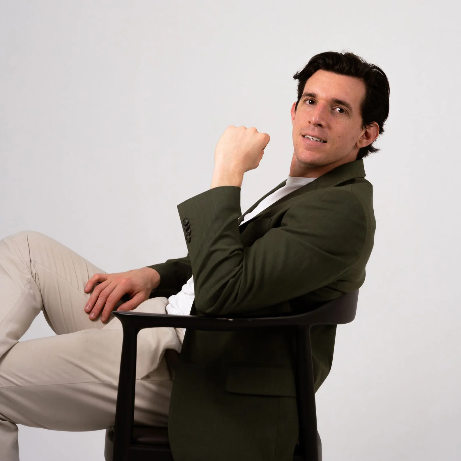 Man sitting in a black chair, wearing a dark blazer and light pants, smiling and flexing his right arm.