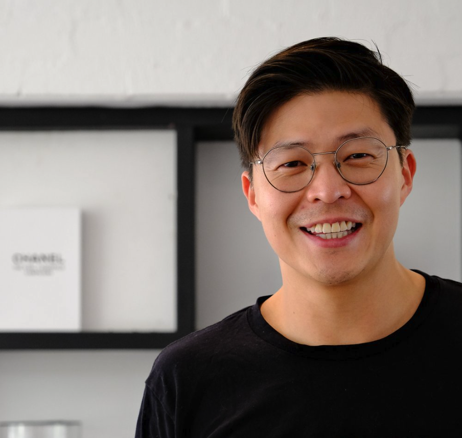 A smiling man with glasses and dark hair, wearing a black shirt, standing in front of a white wall with dark shelving.