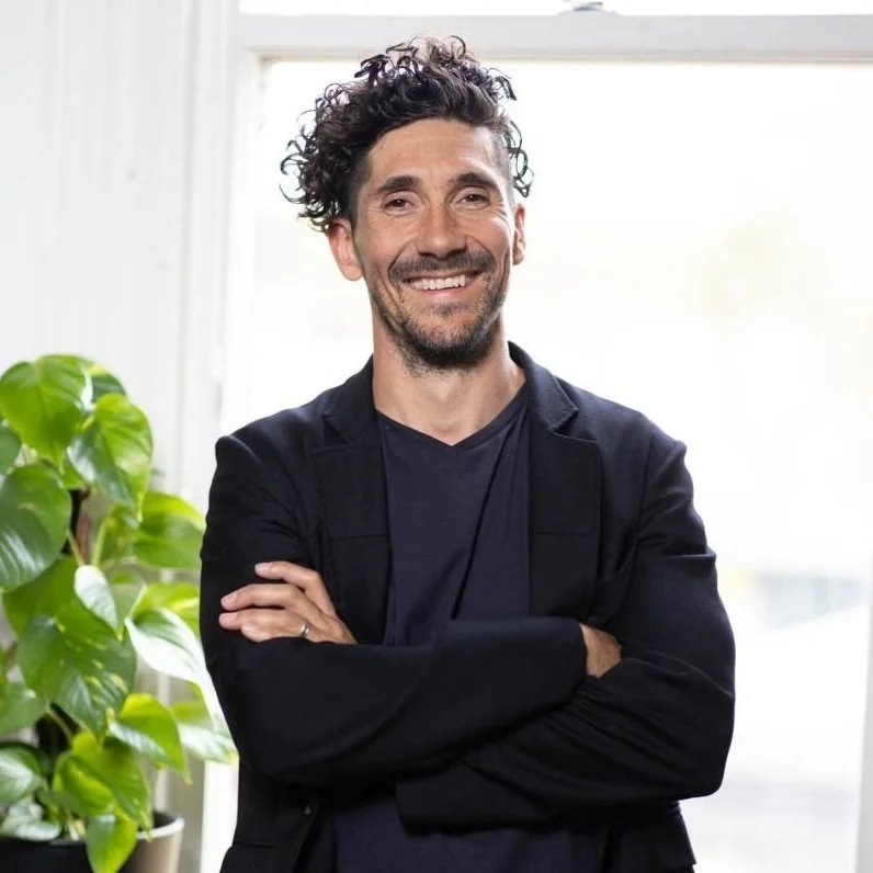 Man with curly hair smiling, standing with arms crossed in front of a window and a green plant inside a room.