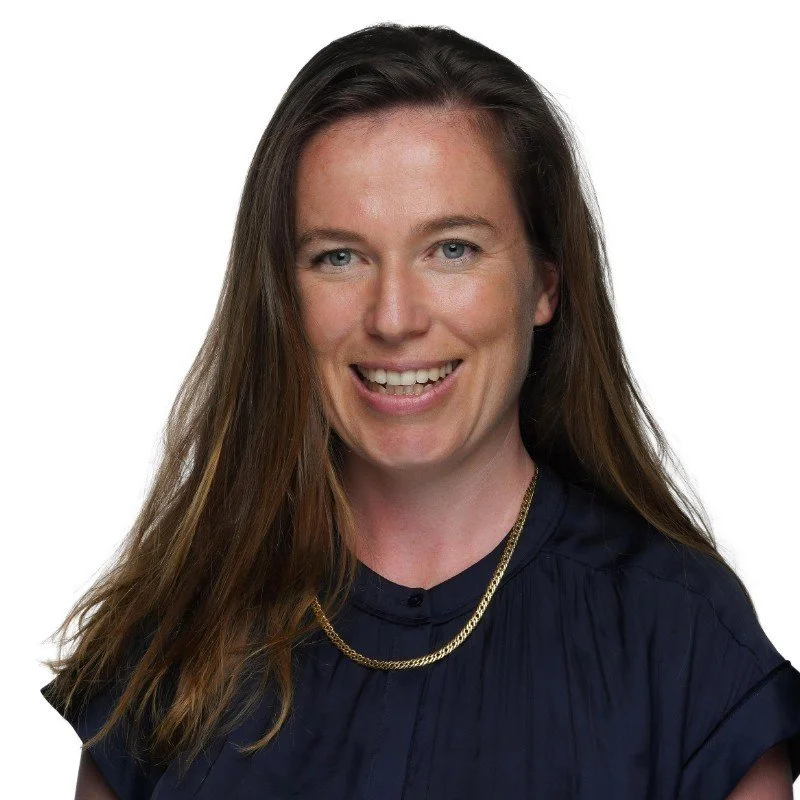 A woman with long brown hair, blue eyes, smiling, wearing a dark blue top and a gold chain necklace.