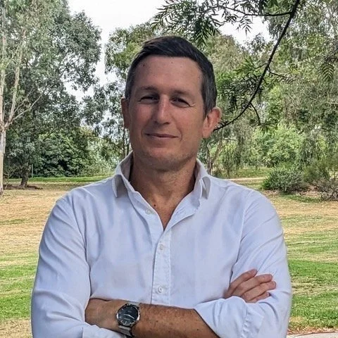A man wearing a white dress shirt with sleeves rolled up, standing outdoors in a park with trees and grass, crossing his arms and smiling.
