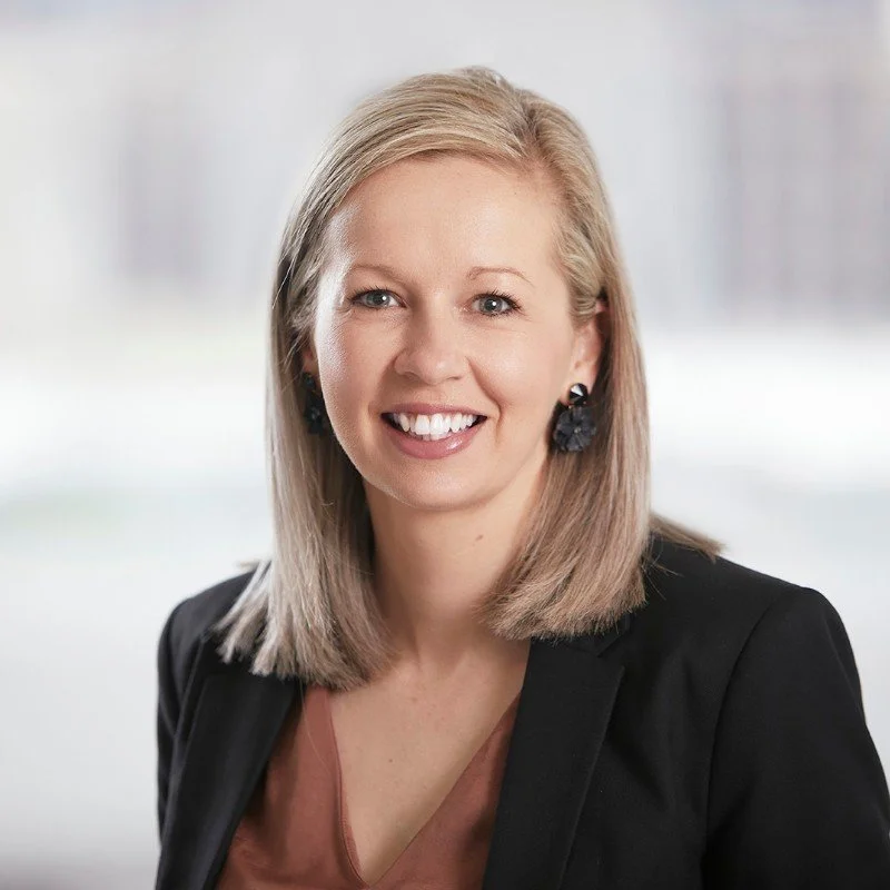 A portrait of a smiling woman with blonde hair, wearing a black blazer, earrings, and a brown top, in front of a blurred out neutral background.