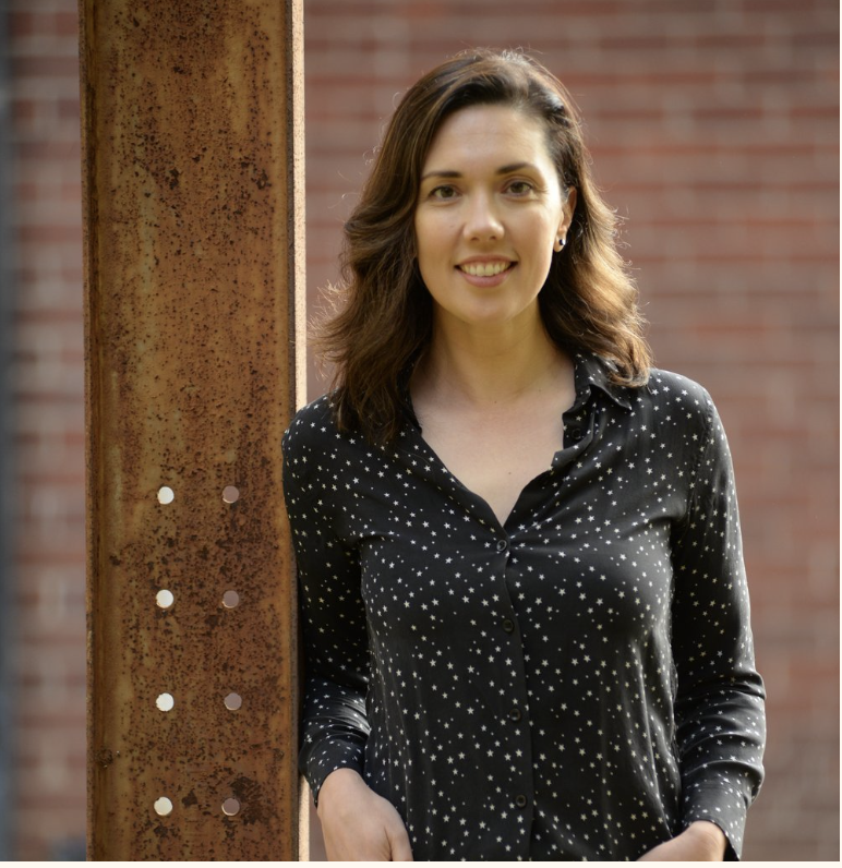 A woman with shoulder-length brown hair smiling, standing outdoors next to a rusty metal post with a brick wall in the background.