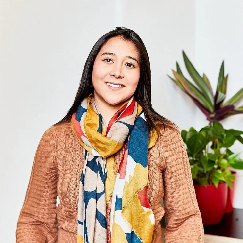 A young woman with long dark hair smiling, wearing a colorful scarf and a brown sweater, standing in front of a potted plant with green and purple leaves.