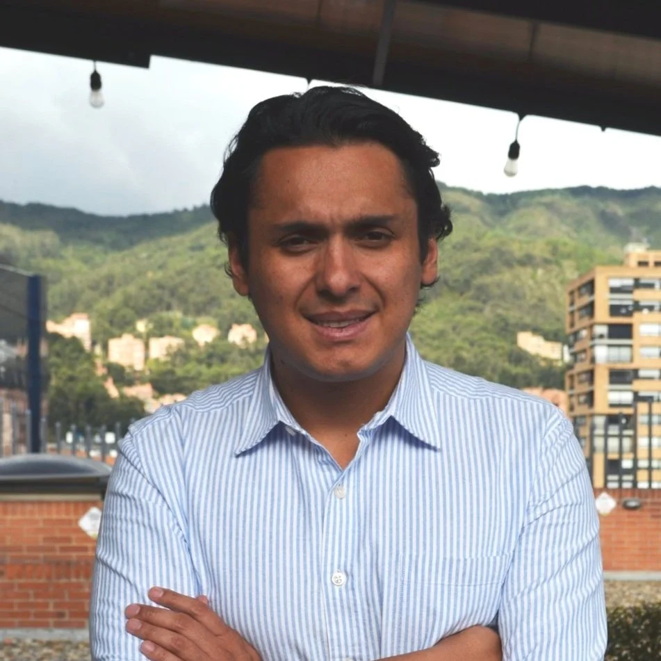 A man with dark hair and a light striped button-up shirt standing outdoors with mountains and buildings in the background.