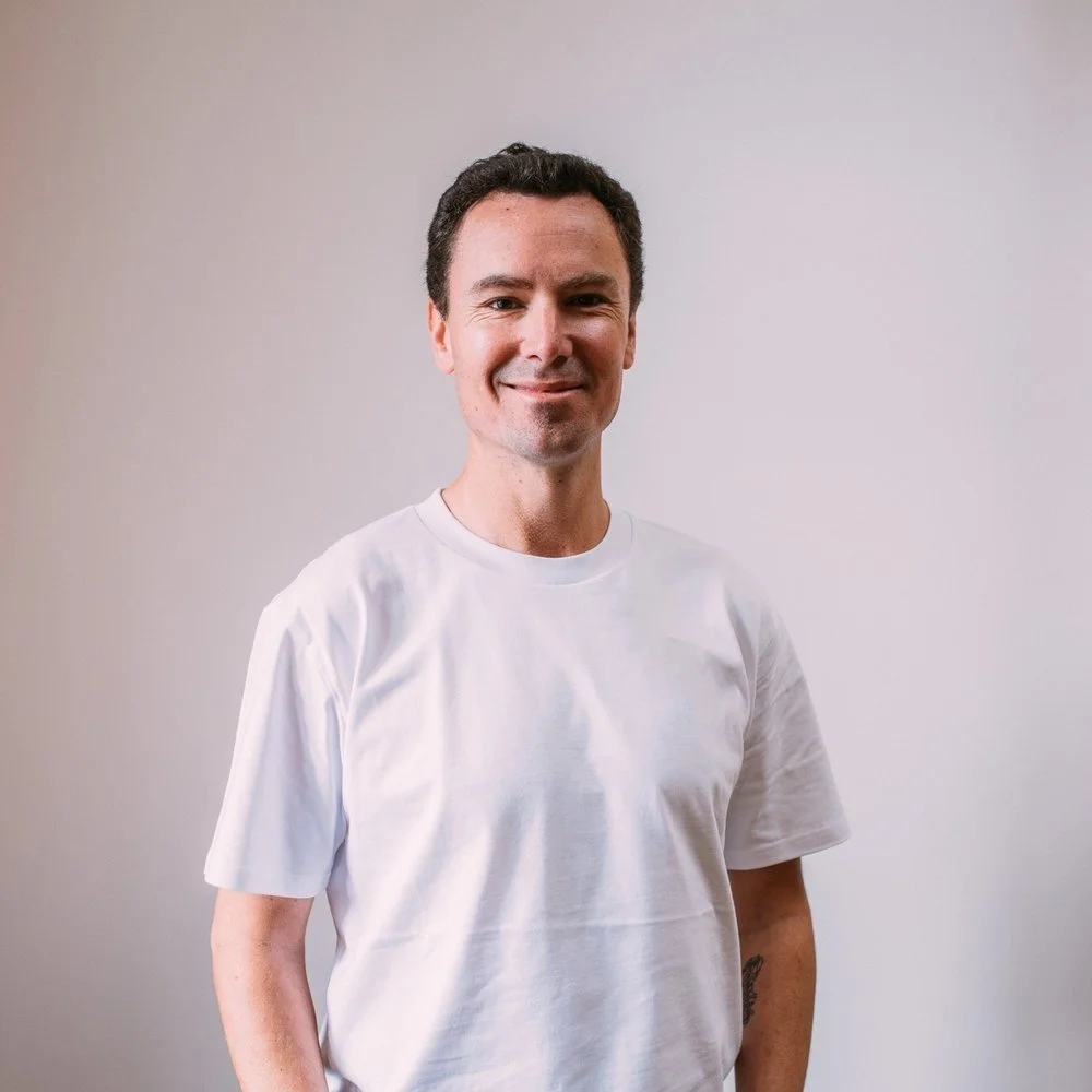A man with dark hair and light skin, wearing a plain white t-shirt, standing against a light-colored wall with a slight smile.