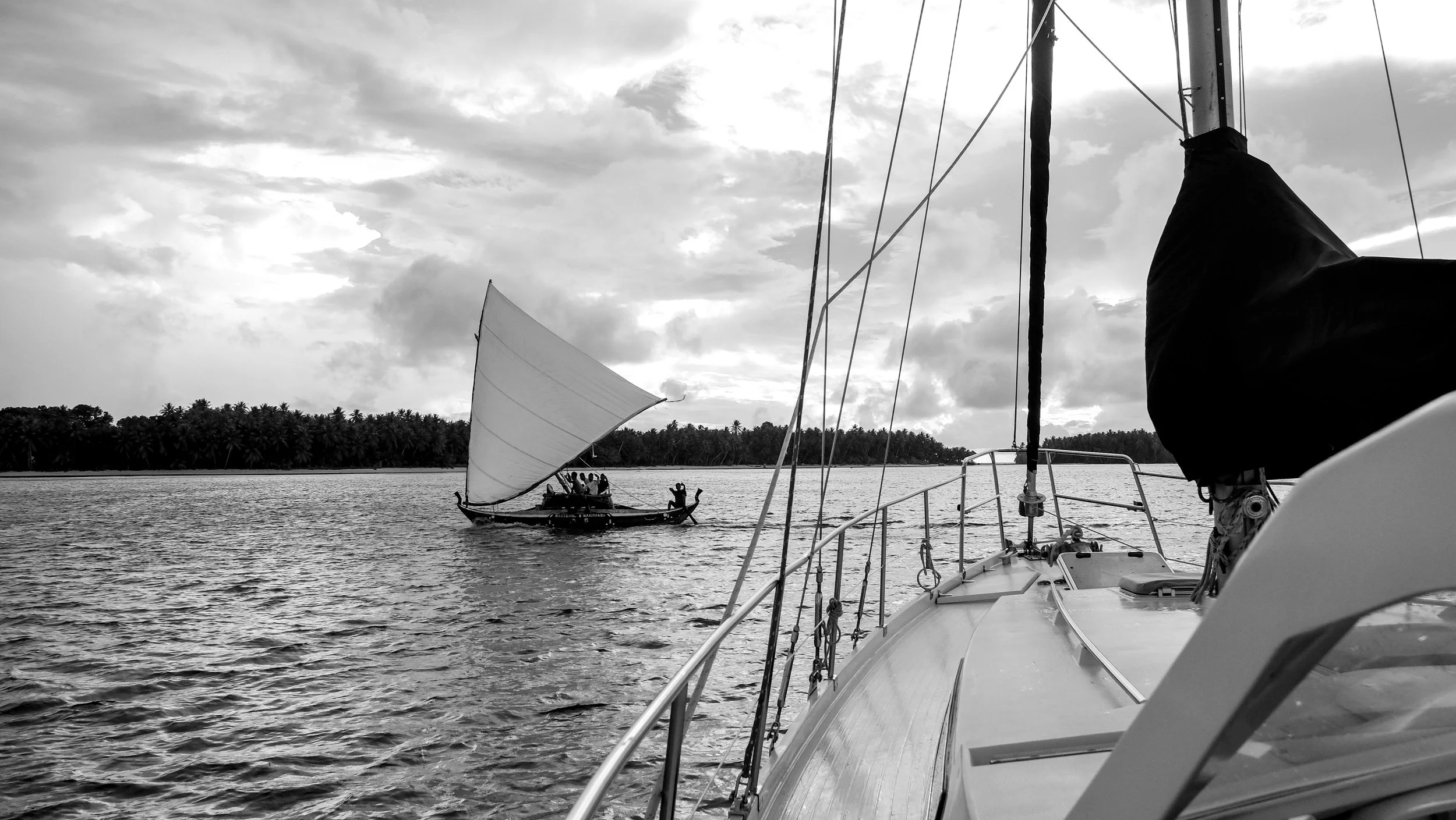 View from a sailboat looking at a traditional wooden boat with a large sail on calm water, with a distant shoreline lined with trees and a cloudy sky in the background.