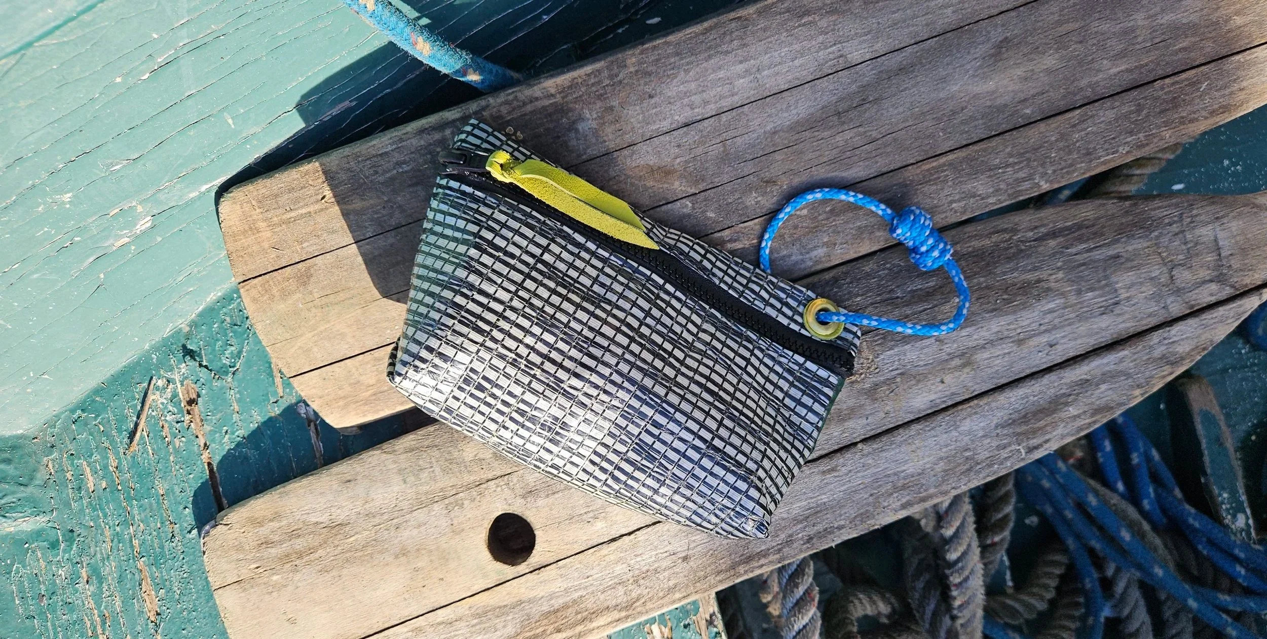 Small black and white checkered pouch with a yellow zipper, scapling on a weathered wooden surface with green paint peeling, near blue ropes.