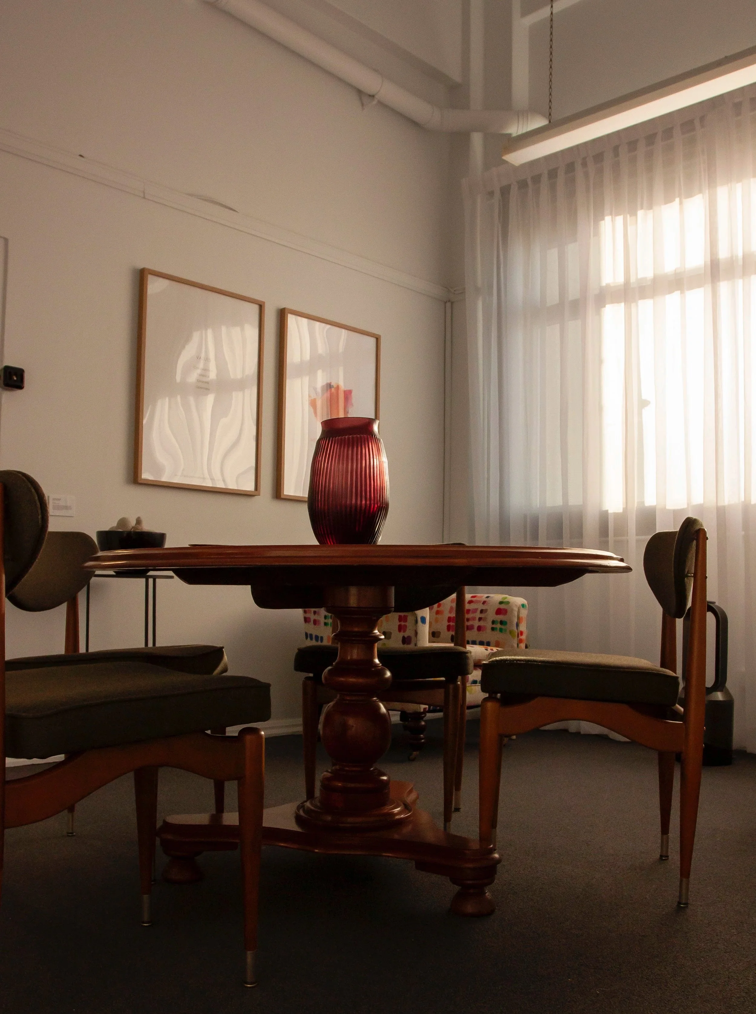An office area with a dark wood table and multiple chairs. Several framed artworks hang on the wall, with sheer curtains covering a large window. A prominent red vase is on the table, with a patterned cushion in the background.