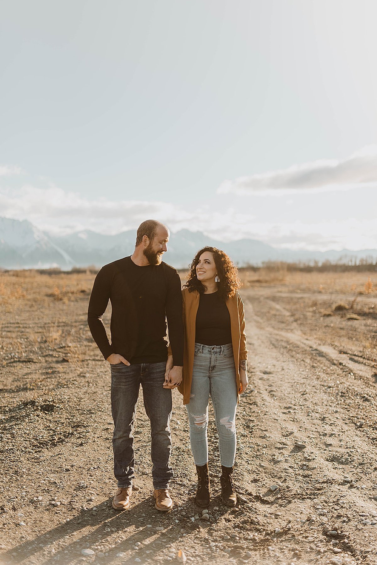  Newly engaged couple stand hand in hand in front of mountain range in shoot by Alaska wedding photographer 