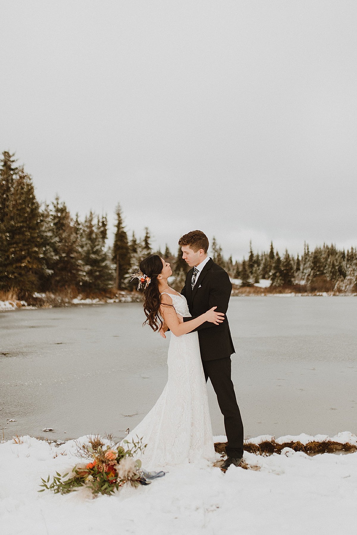  bride and groom pose at winter lake with pine trees and autumn bouquet in snowy fall wedding  