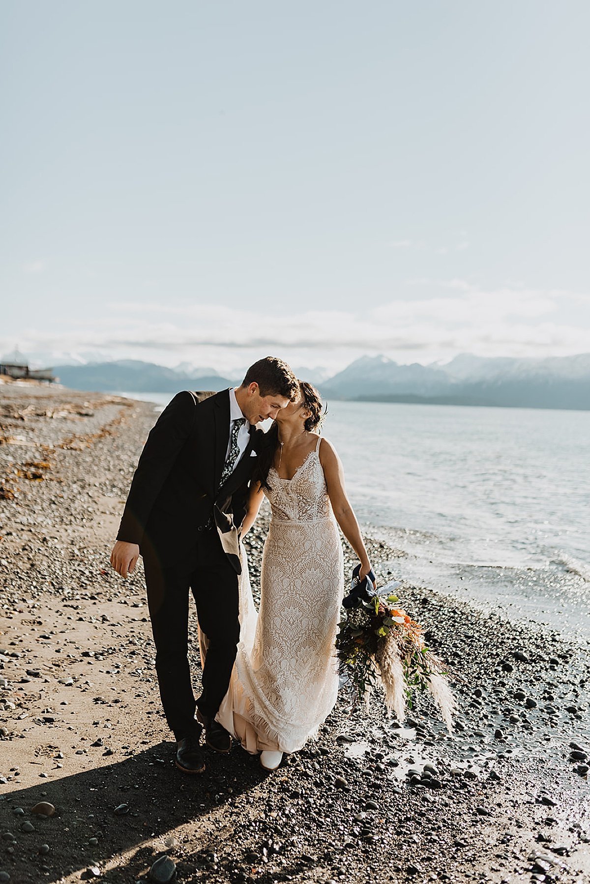  bride and groom walk along rocky lake beach before snowy fall wedding 
