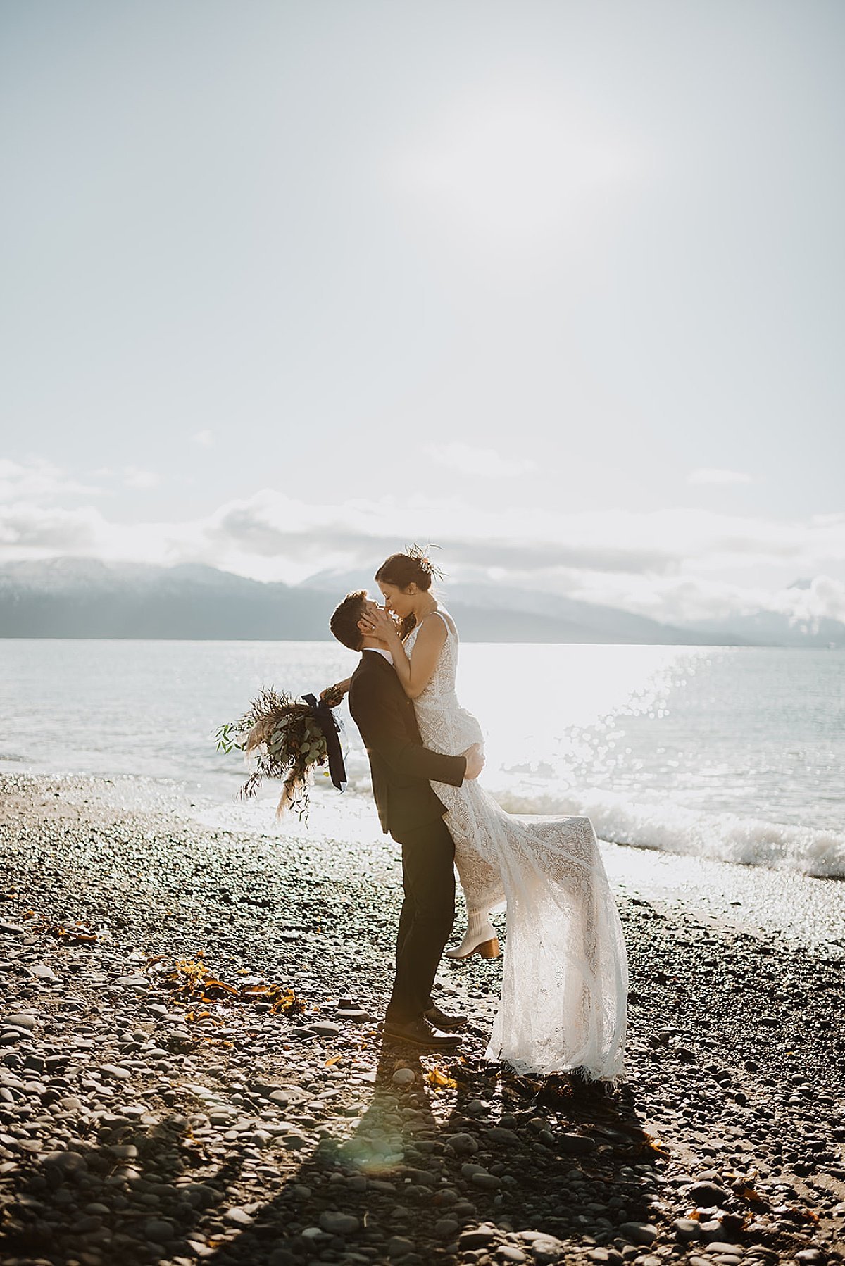  bride and groom pose with a kiss on mountain lake beach at ceremony shot by alaska wedding photographer 