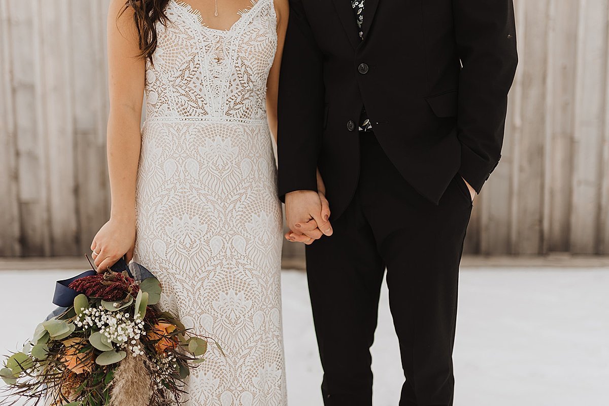  bride in white lace gown and groom in tuxedo pose with autumn color bouquet during snowy fall wedding 