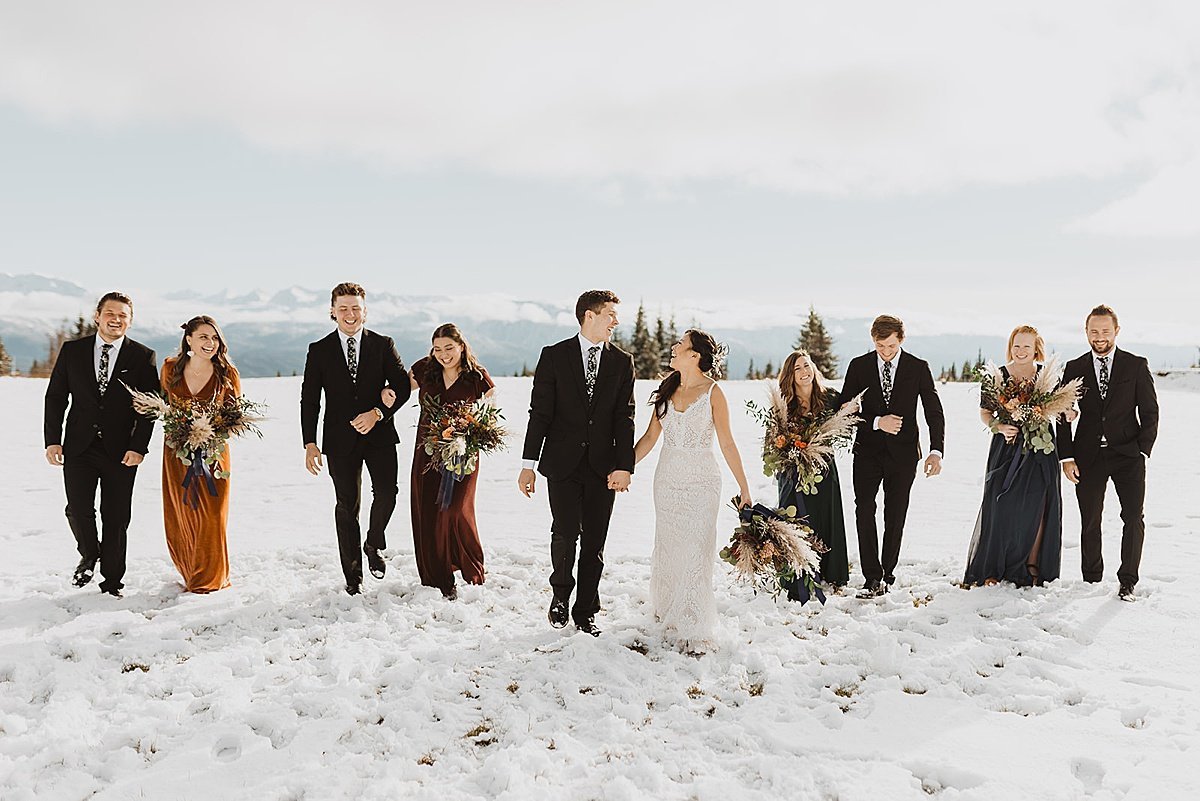  bride, groom, and wedding party wearing autumn colors walk across field during snowy fall wedding 