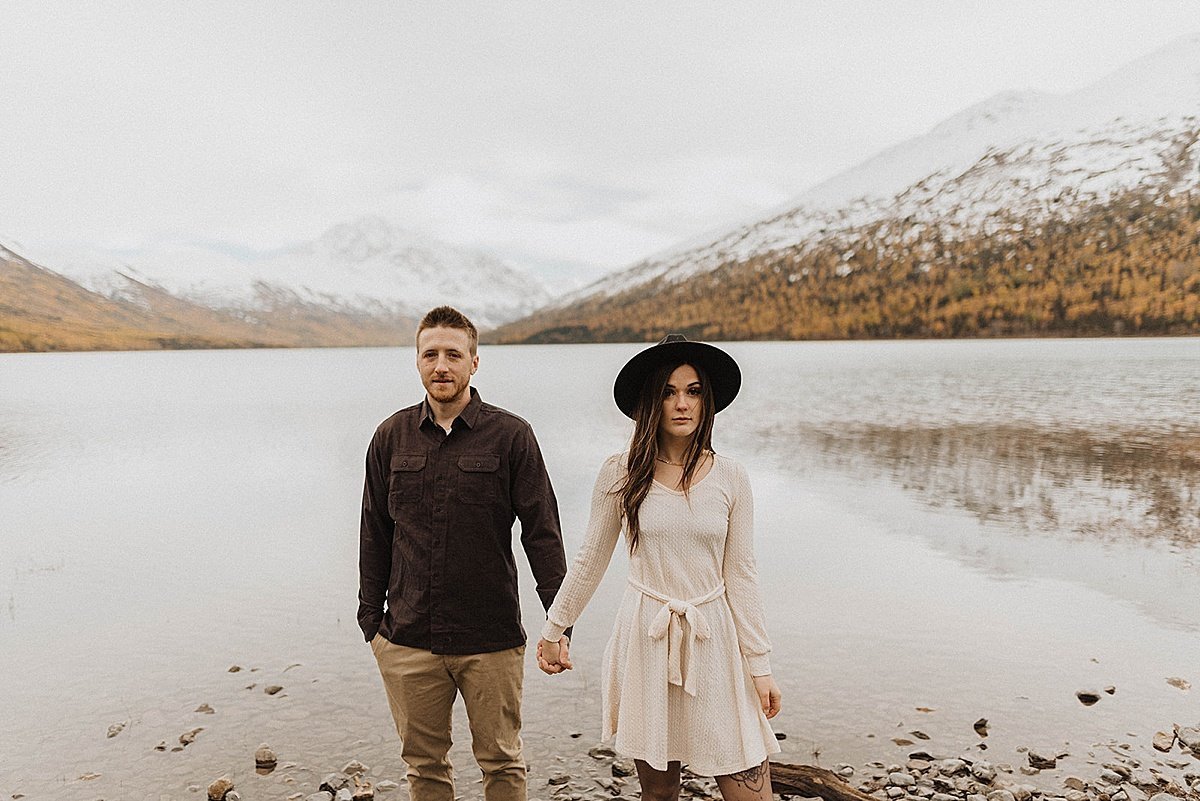  couple stand hand in hand in front of moody lake in the mountains during outdoor engagement shoot by alaska wedding photographer 