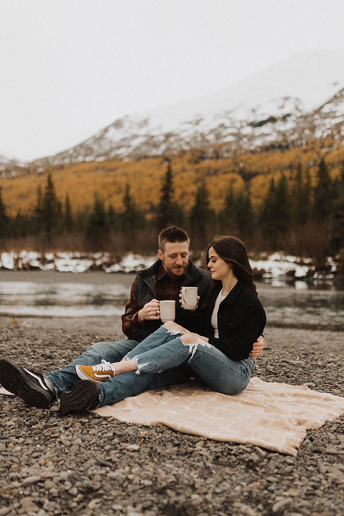 engaged couple hold mugs and enjoy coffee while sitting on a blanket at mountain lake during autumn engagement shoot 