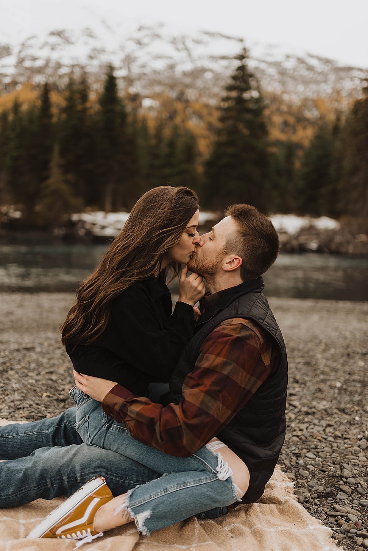  cute couple in jeans and plaid flannel and sneakers kiss on blanket at outdoor engagement shoot by alaska wedding photographer 