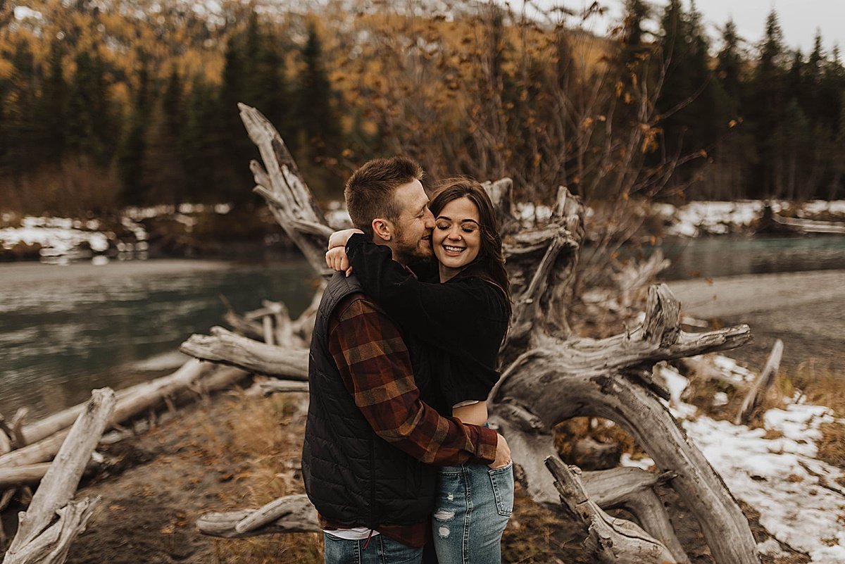  newly engaged couple in flannel jackets kiss in front of rugged tree at mountain lake shoot by theresa mcdonald 