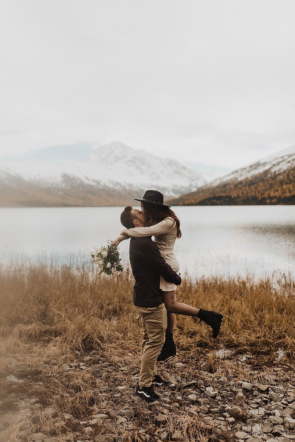  couple wearing boho mountain chic clothing kiss in front of mountain lake during autumn engagement shoot 