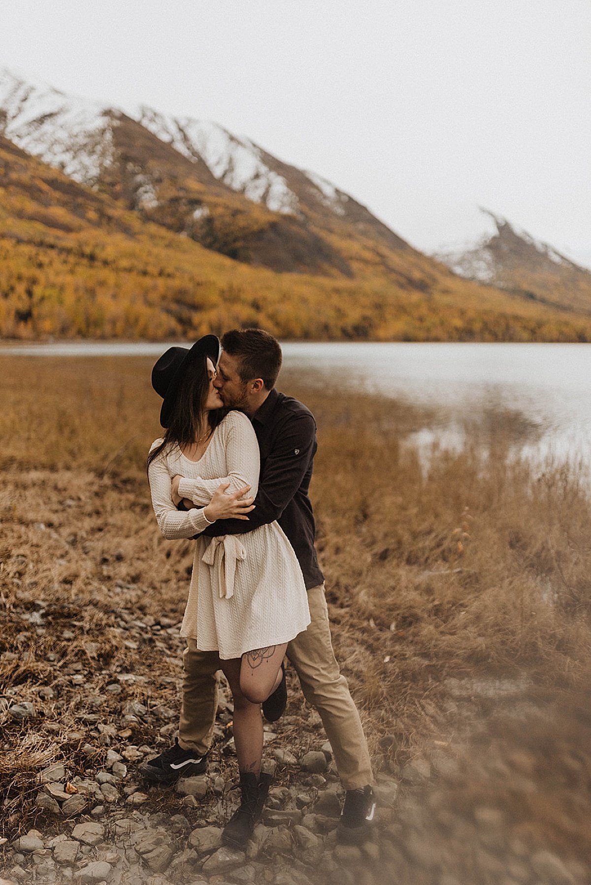  man and woman in boho felt hat and cute dress kiss by mountain lake in fall engagement shoot by alaska wedding photographer 
