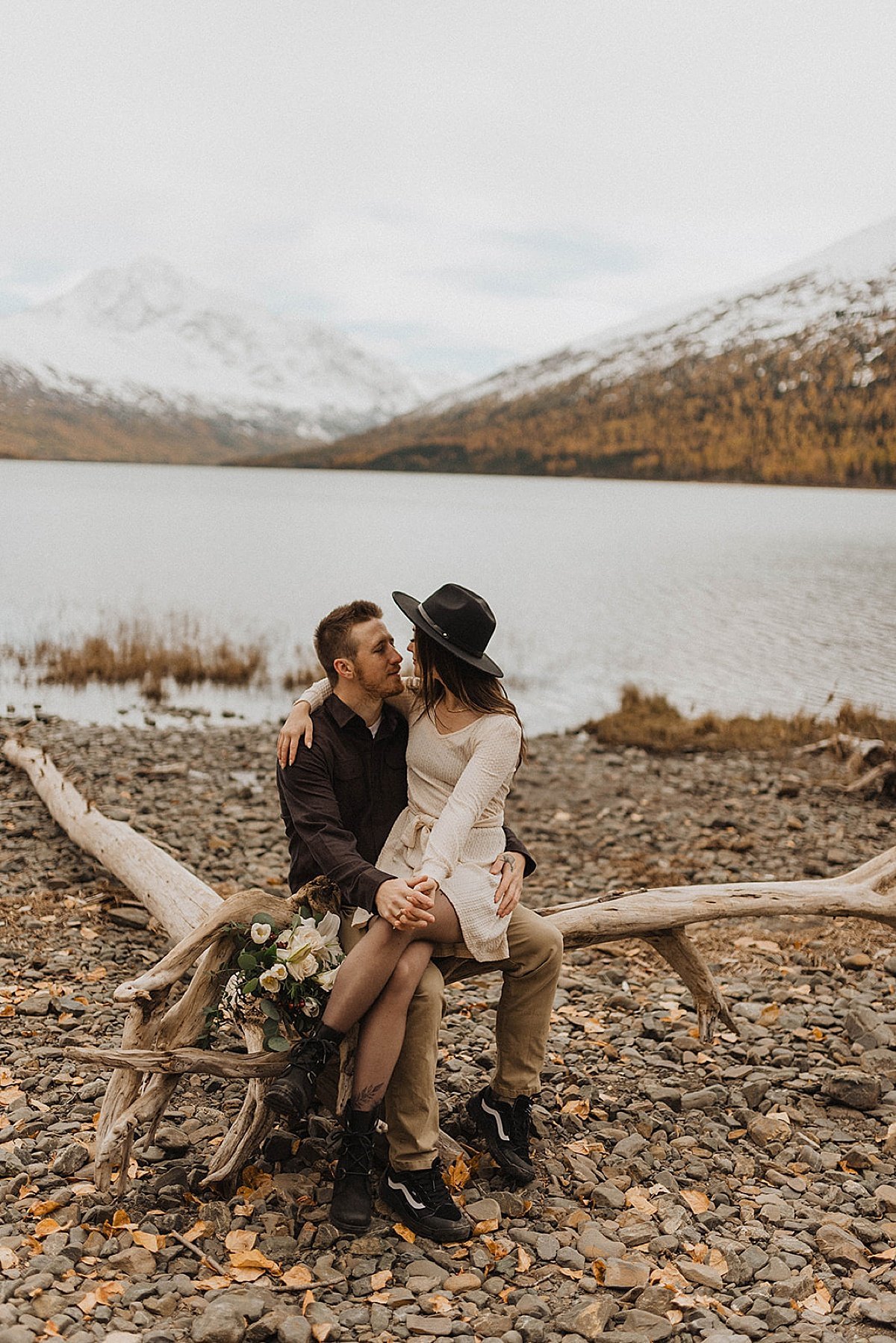  man and woman in boho felt hat embrace with wildflower bouquet on rocky mountain lake beach in autumn engagement shoot 