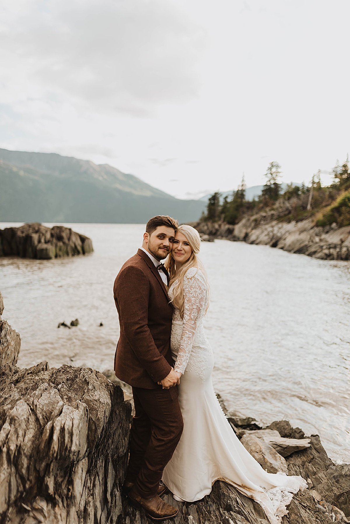  newlywed couple pose at mountain lake in burgundy suit and white lace gown after moody glacier creek wedding 