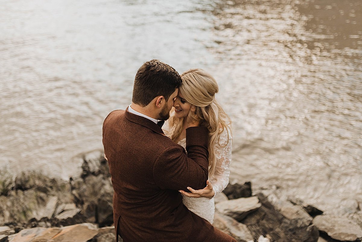  bride in white lace gown and groom in burgundy suit embrace on rocky lakeshore at shoot by alaska wedding photographer 