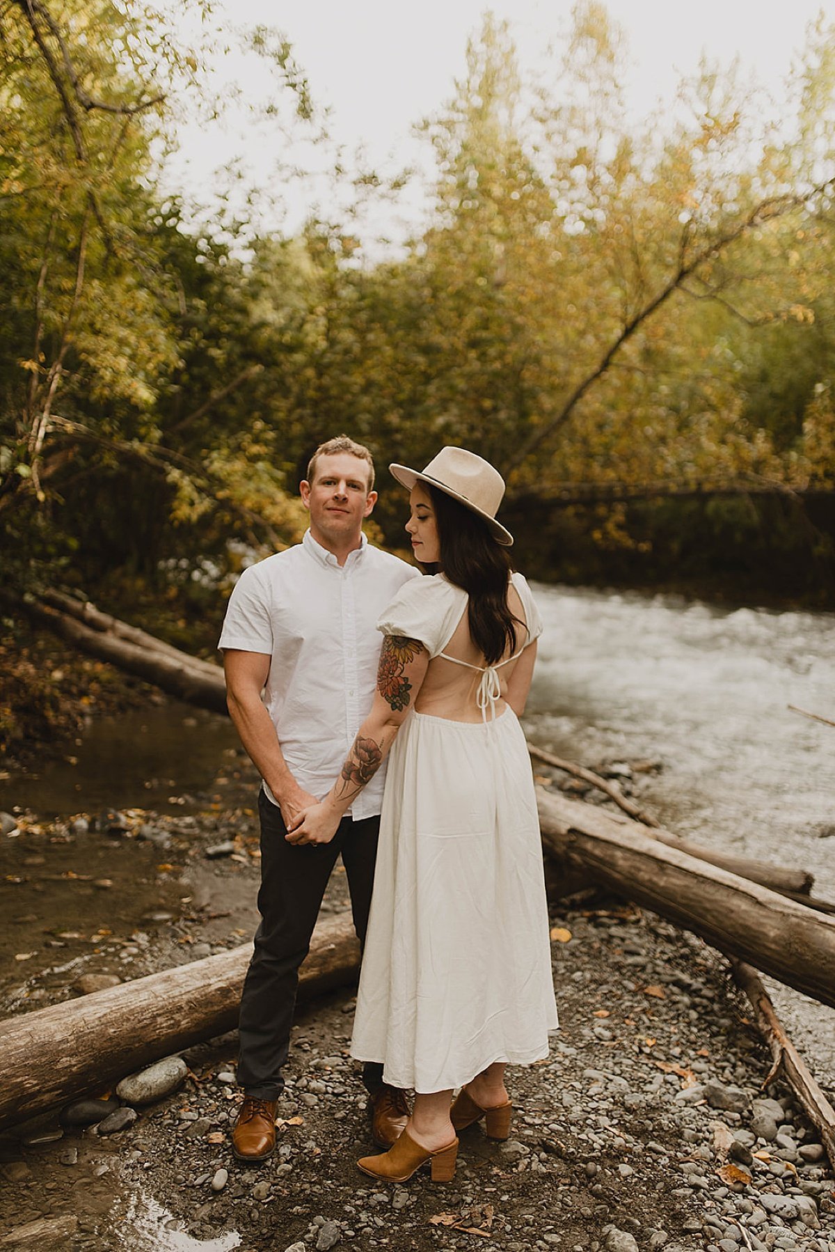  Couple stand hand in hand by river in woodsy engagement shoot 