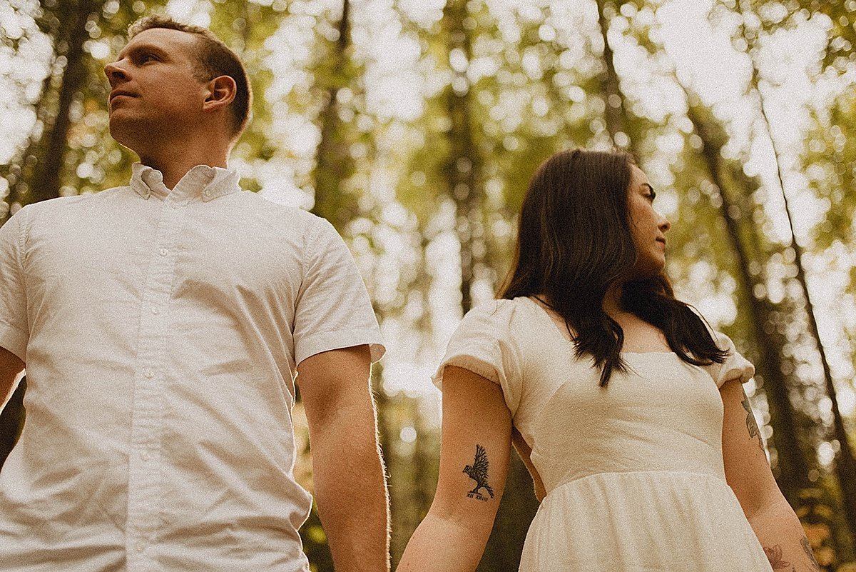  Couple in white shirt and dress stand hand in hand for woodsy engagement shoot 